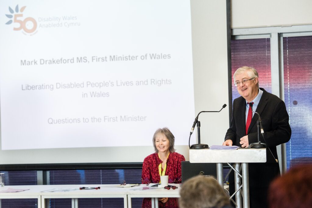 Wales First Minister, Mark Drakeford, standing behind a lectern. His name is on a big screen behind him above the words 'Liberating disabled people's lives and rights in Wales.' DW Chief Executive, Rhian Davies, sits behind a table to the First Minister's right. They're both smiling.