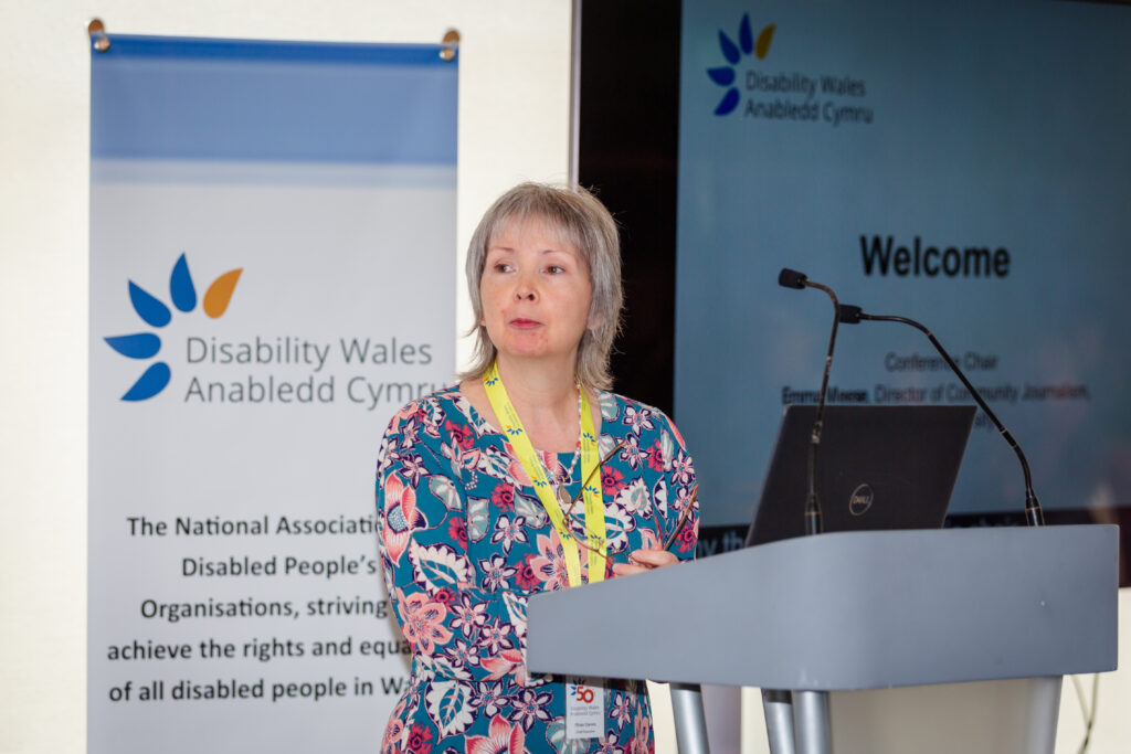 Rhian Davies standing behind a podium which has multiple microphones, delivering her speech. She has short grey/blonde hair and wears a colourful patterned top and a Disability Wales lanyard. Behind her is a screen with DW's logo on it.