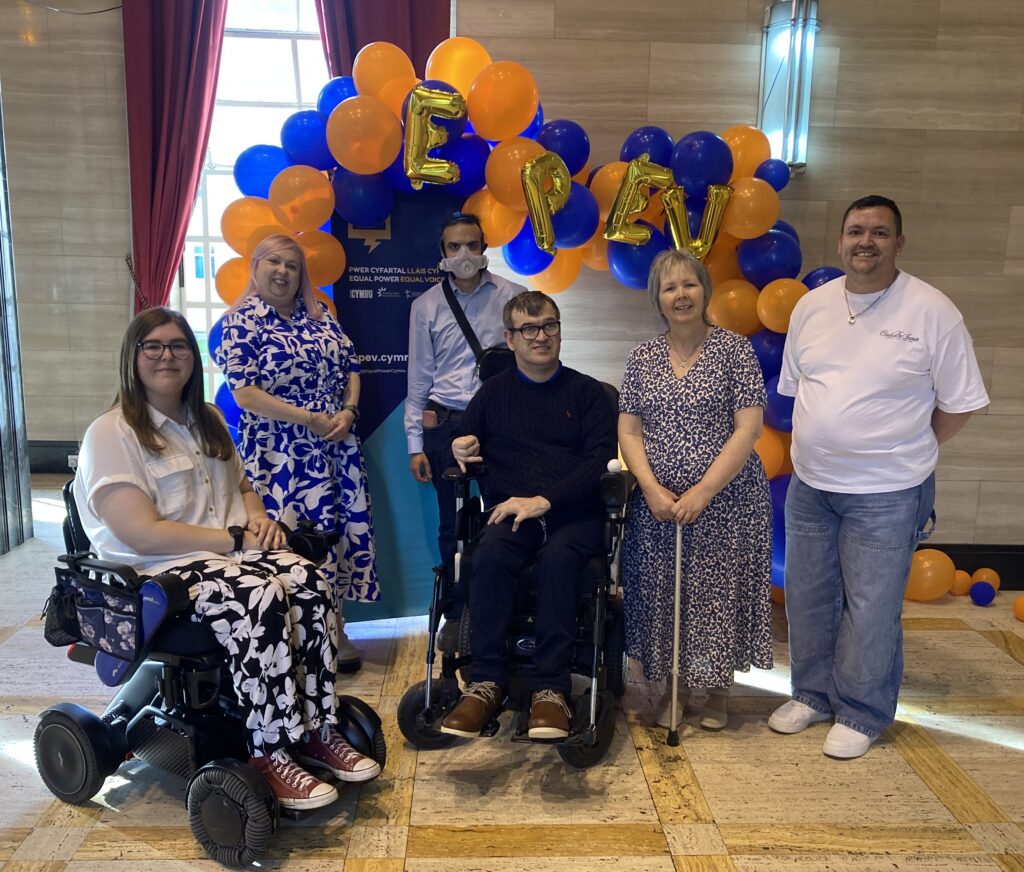 DW Chief Executive, Rhian Davies, and EPEV Programme Coordinator, Linus Harrison, with mentees in front of an orange and blue balloon arch with EPEV picked out in gold lettered balloons. Everyone is smiling at the camera, delighted to be celebrating their achievements as part of Equal Power Equal Voice.