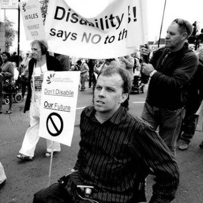A black and white photo of Simon Green holding a sign that says Don't disable our future