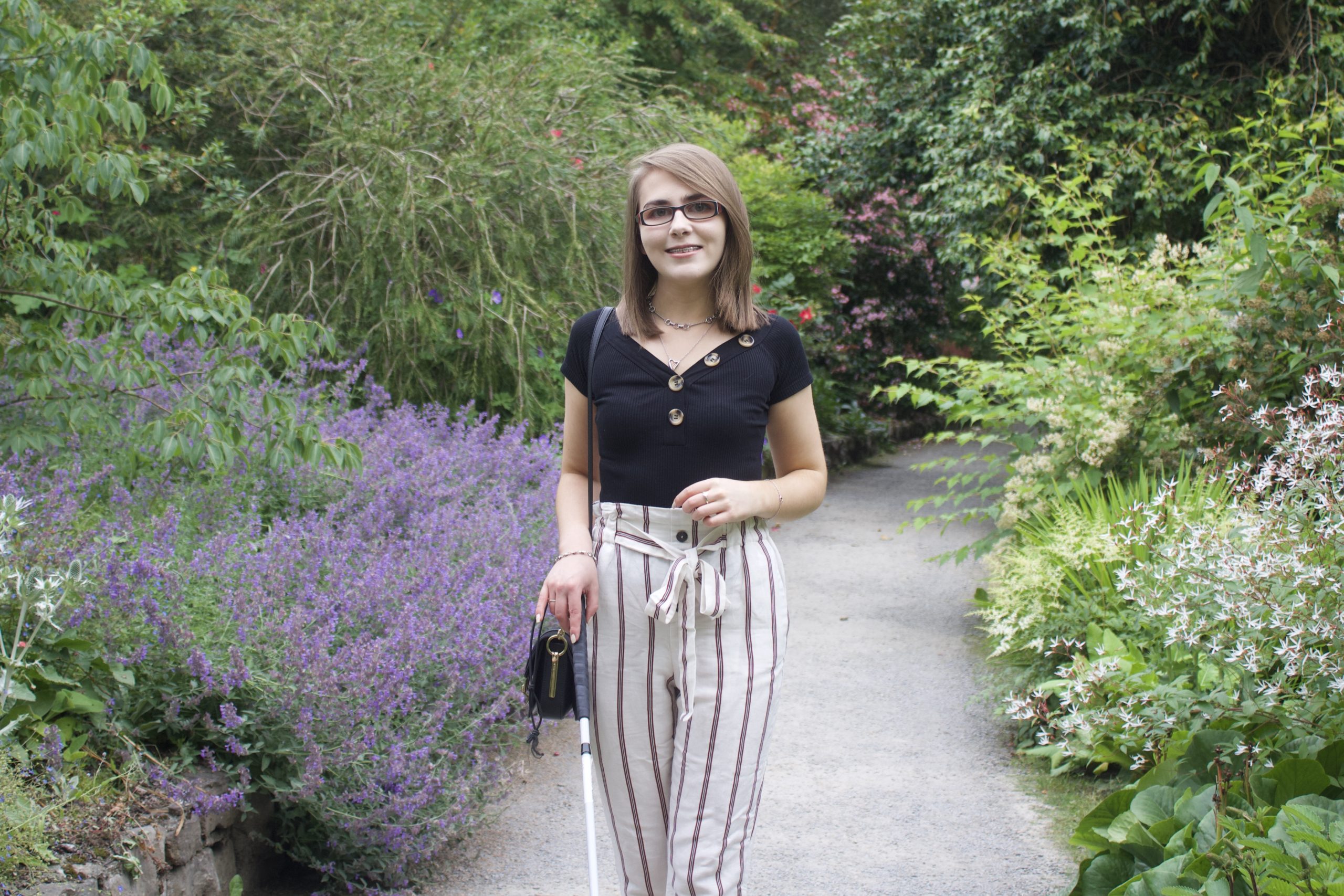 A photo of Elin Williams - a woman with a black top and striped trousers stands on a path surrounded by greenery