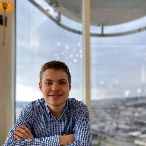 Alex sitting by a table in a restaurant with a backdrop of Swansea City Centre on an overcast autumn day. His arms are crossed and rest on the table.