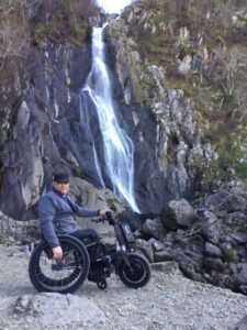 John on his hand bike in front of a waterfall amongst rocky surroundings