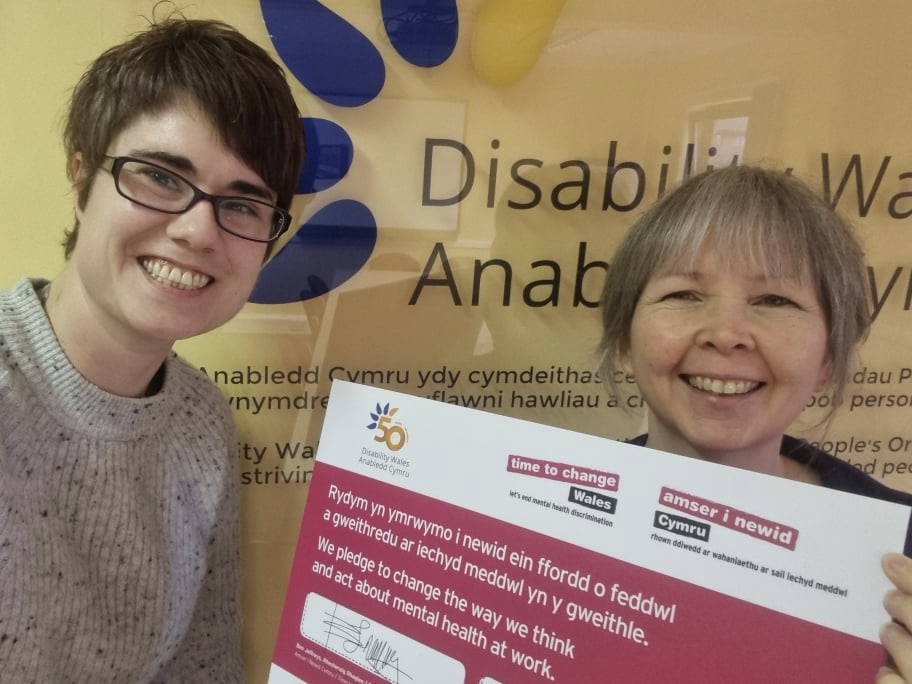 Disability Wales Chief Executive, Rhian Davies, and Information Officer, Alex Osborne, holding the Time to Change Wales pledge which has white text on a red background. Both women smile widely at the camera and stand in front of a wall which shows the Disability Wales logo.