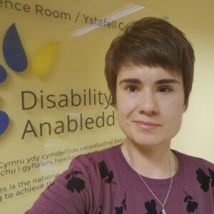 Alex, a white woman with short brown hair, stands in front of a wall which has the Disability Wales logo on it. She wears a mauve coloured top with a dainty silver necklace.