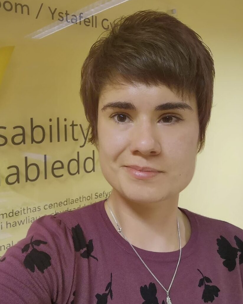Alex, a white woman with short brown hair, stands in front of a wall which has the Disability Wales logo on it. She wears a mauve coloured top with a dainty silver necklace.