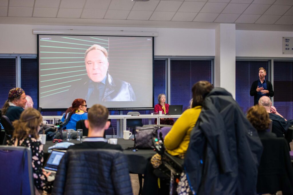 Mik Scarlet on-screen addressing a room full of people who sit around round tables.