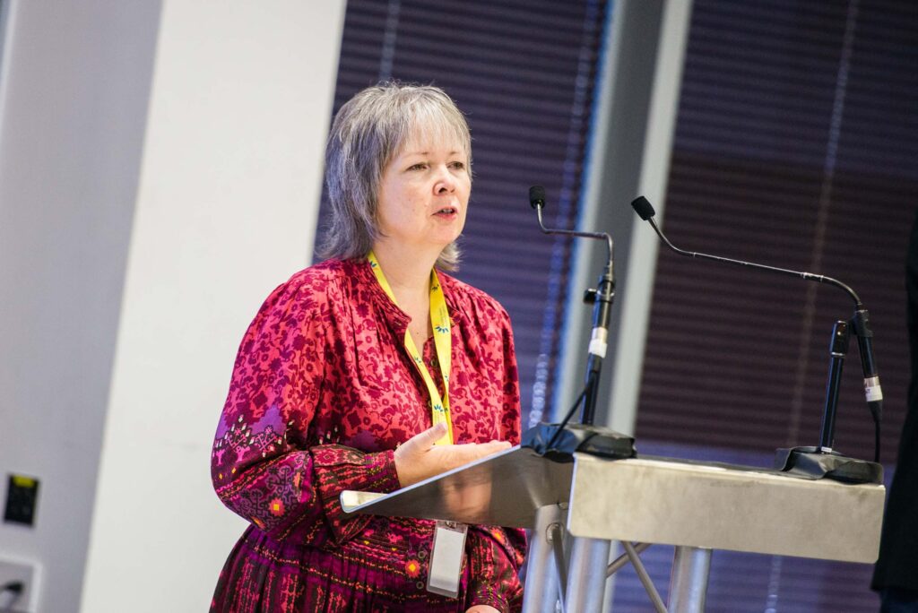 Rhian Davies standing at a lectern. She has short light hair and is wearing a pink floral top with a DW lanyard around her neck.