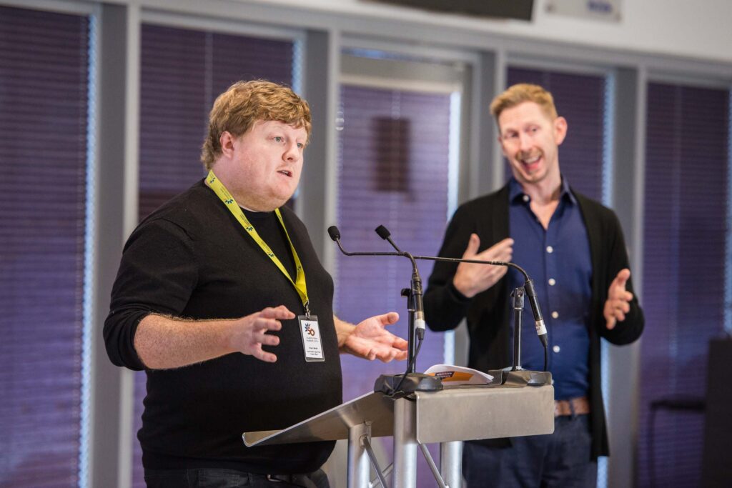 Equal Power Equal Voice Mentoring Project Officer, Shaun Bendle, standing behind a lectern. He wears a black jumper and a DW lanyard. A BSL interpreter is seen signing to his left.