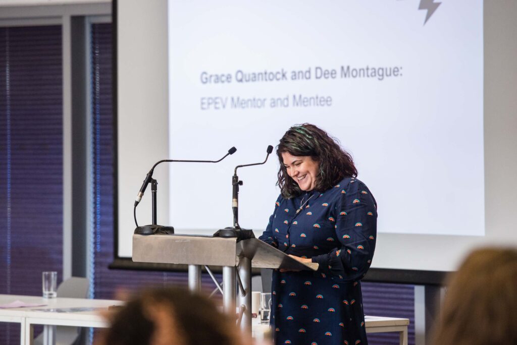 Equal Power Equal Voice mentee, Dee Montague, standing at a lectern delivering a speech.She's smiling as she looks at her notes. A big screen is seen behind her with Grace Quantock and Dee's names on it above words that read EPEV Mentor and Mentee.