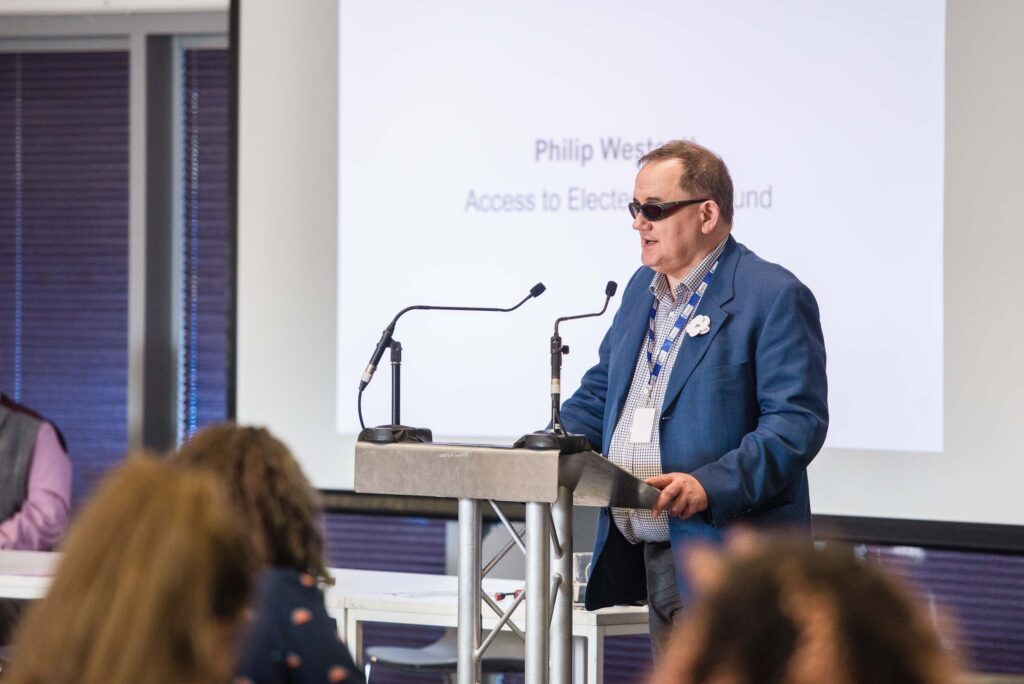 DW's Civic Participation Officer, Philip Westcott, standing at a lectern with his hands holding either side of it. A big screen behind him shows his name above the words Access to Elected Office Fund.