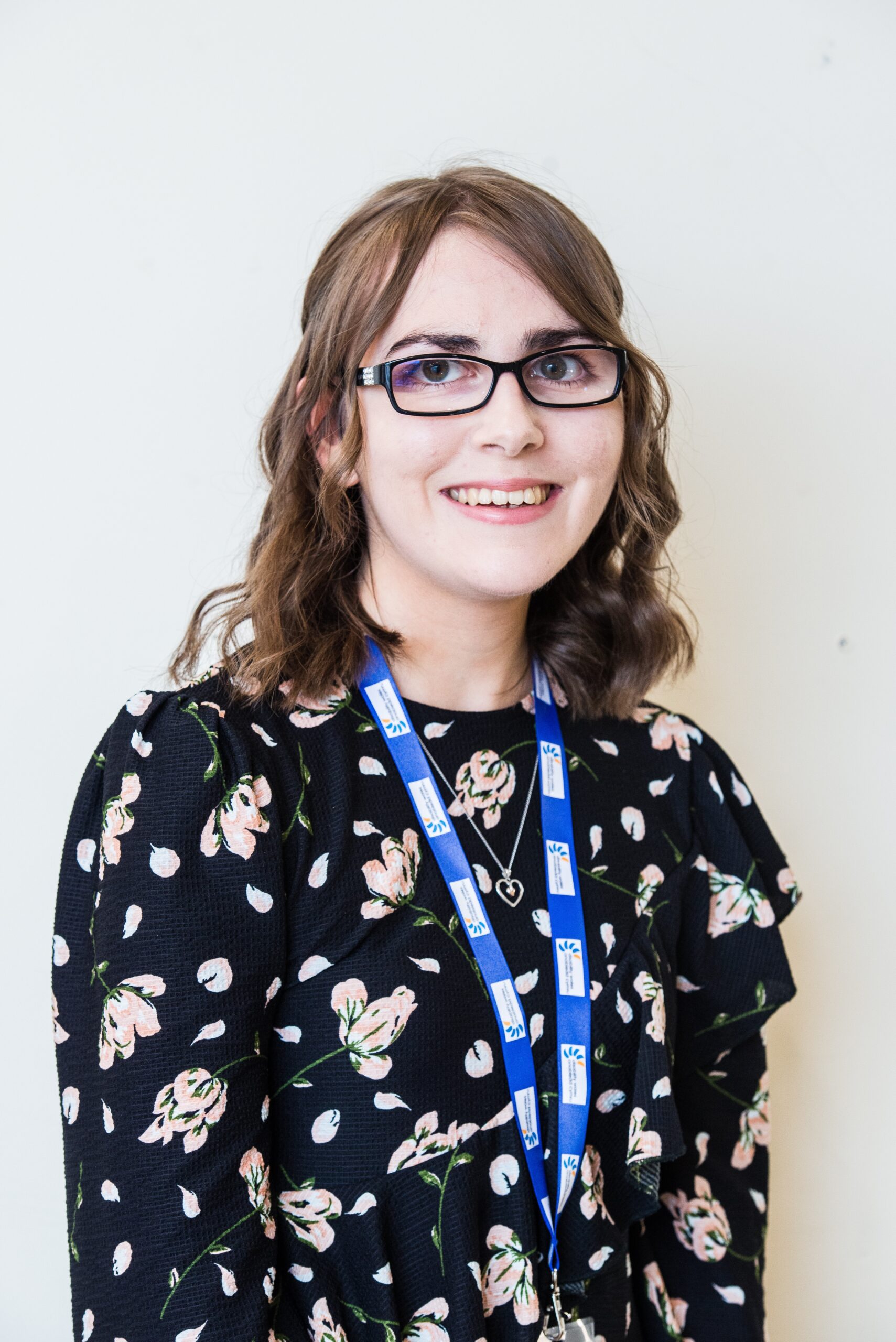 Elin Williams standing in front of a white wall and smiling at the camera. She wears a black floral dress along with a DW lanyard and black glasses with silver crystals on the side. Her shoulder length brown hair is curled and styled into a half-up, half-down do.