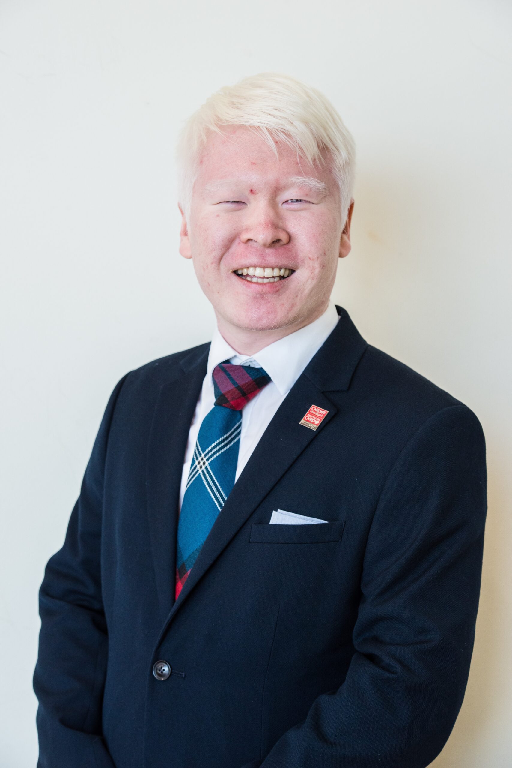 Board member, Felix, standing in front of a plain white wall and smiling widely at the camera. He wears a black suit with a white shirt and blue tie. A small handkerchief peeks out from the top of his left suit jacket pocket.