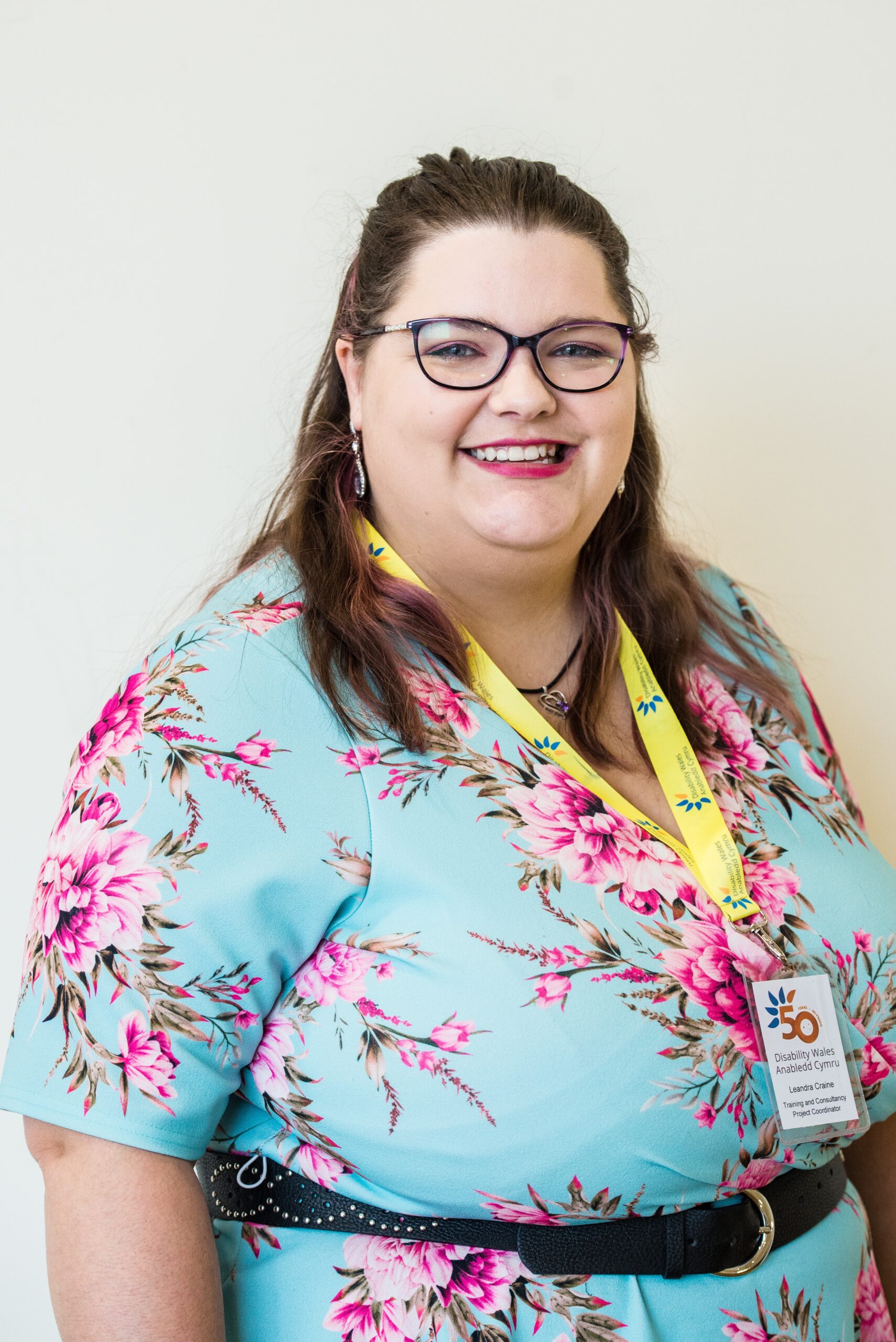 Leandra Craine standing in front of a white wall and smiling at the camera. She wears a turquoise dress with pink flowers on it along with a DW lanyard and glasses. Her dark hair is styled into a half-up, half-down do.