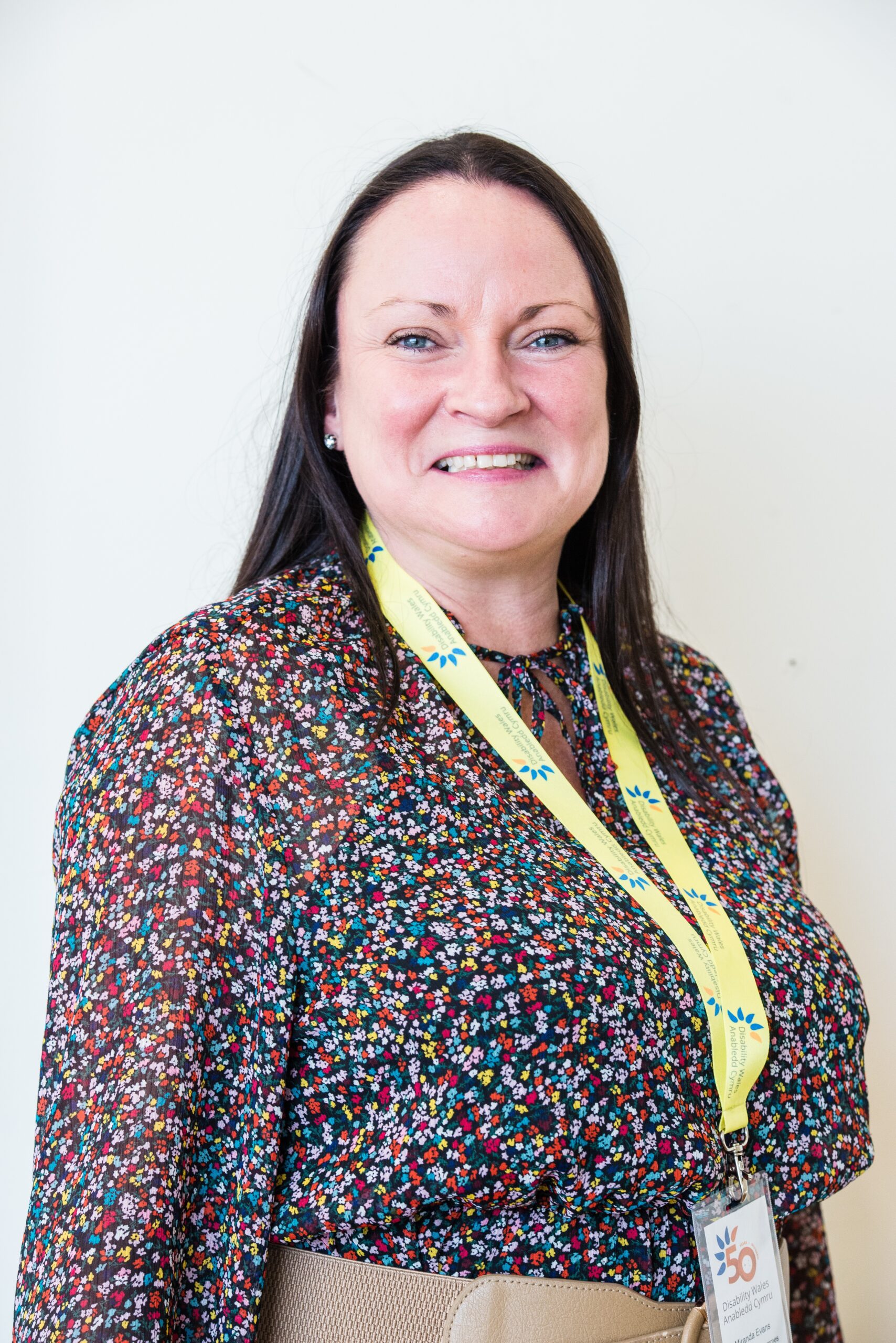 Miranda Evans standing in front of a white wall and smiling at the camera. She has long brown hair and is wearing a multicoloured patterned blouse with a DW lanyard hanging from her neck.