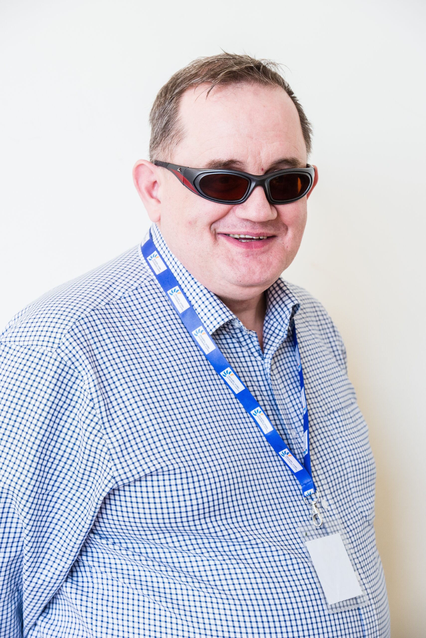 Philip Westcott smiling at the camera. He wears a blue check shirt, a DW lanyard and sunglasses. He has brown hair.