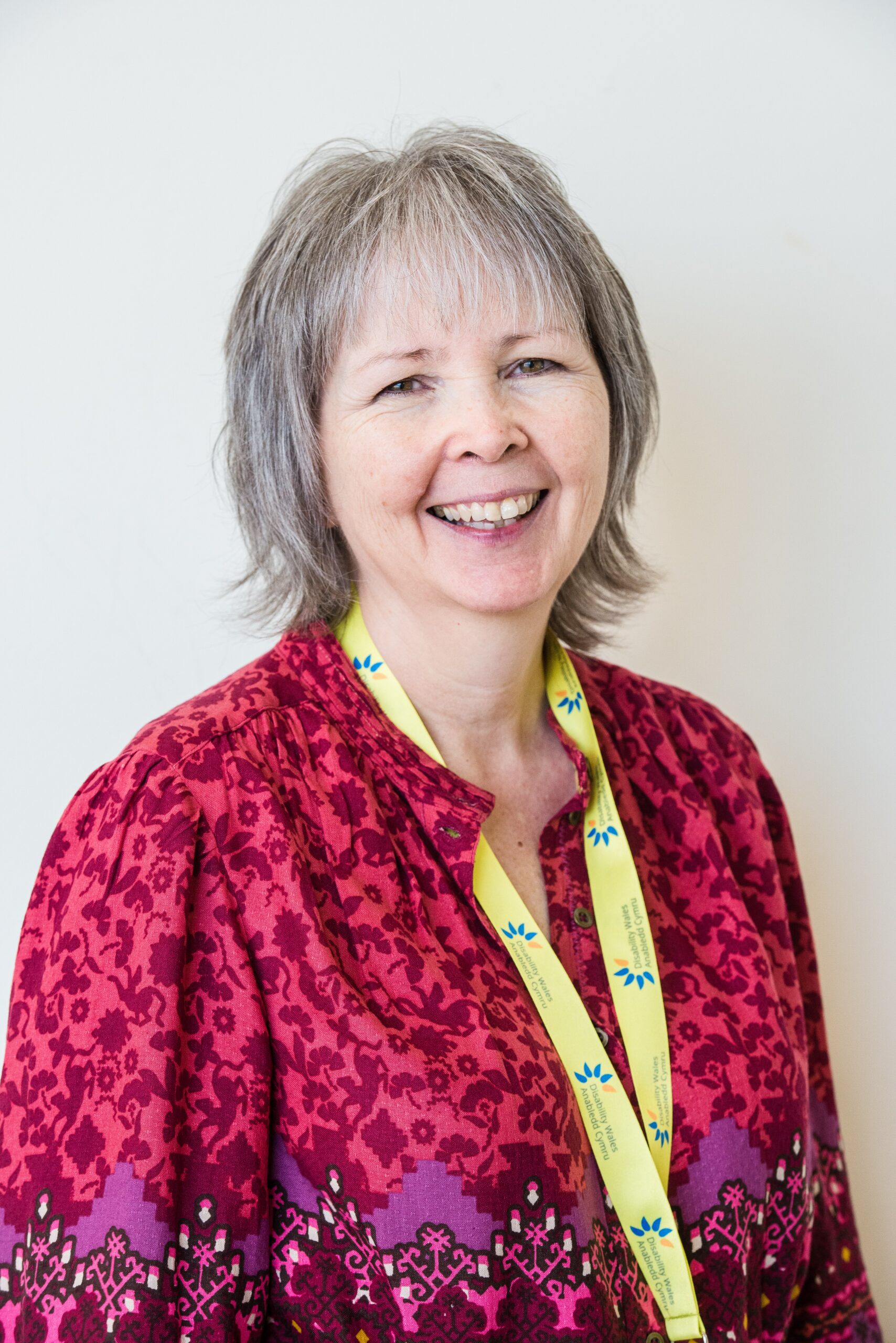 DW Chief Executive, Rhian Davies, standing in front of a white wall and smiling broadly at the camera. She has light short hair and is wearing a bright pink floral top. A Disability Wales lanyard hangs from her neck.