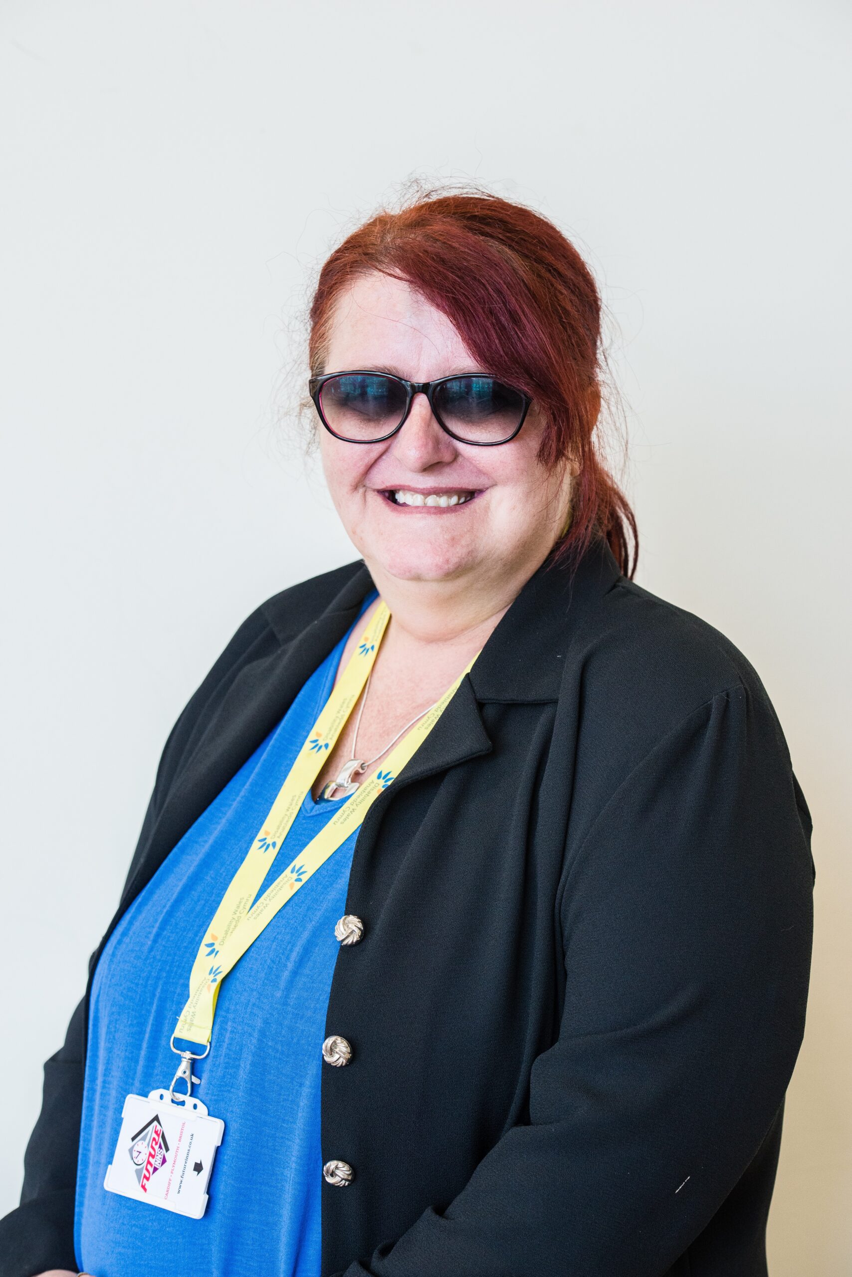 Chairperson, Willow, standing in front of a white wall. She wears a dark blazer over a bright blue top. Her raven hair is tied back and she's smiling at the camera.