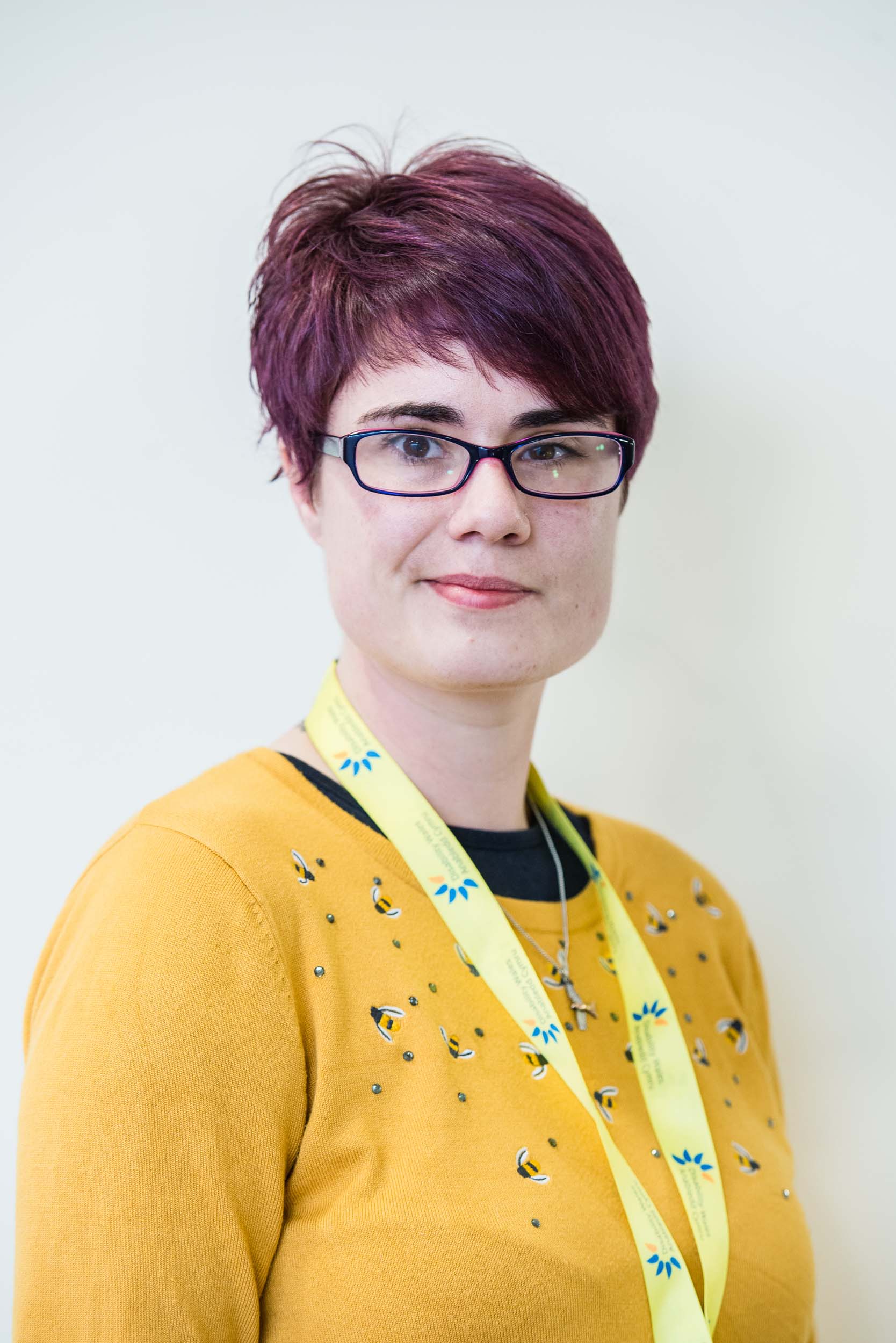 Alex Osborne standing in front of a white wall. She has short purple hair and wears a bright yellow jumper. A DW lanyard is worn around her neck.