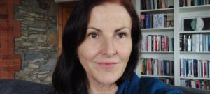 A headshot of Professor Debbie Foster, a white woman with long dark hair and brown eyes. There is a big white bookcase behind her displaying a variety of books.