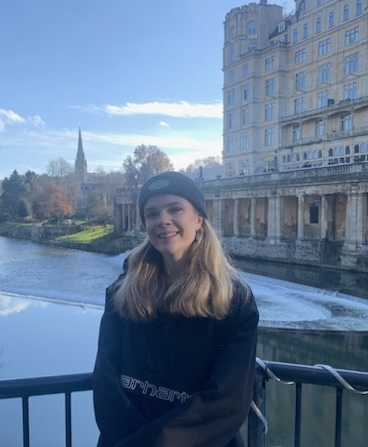 On a hazy day, Rosie, a white woman with long blonde hair, stands against a railing in front of a body of water with buildings in the background. She wears a dark outfit with a beanie hat and is smiling at the camera.