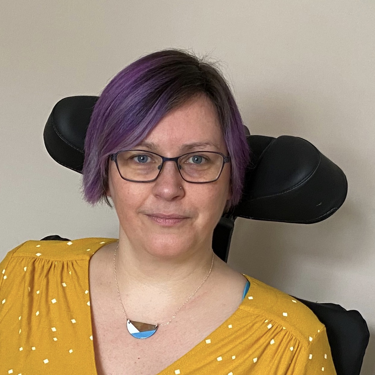 A head and shoulders shot of Dr Aimee Grant who sits in front of a plain light coloured wall. She has short purple hair and is wearing glasses. She is smiling slightly and looking directly at the camera. She is wearing a mustard yellow top with white polka dots and a necklace in a geometric pattern featuring blue, white and black colours.