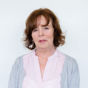 Tracey Blockwell in front of a plain white wall. She has short, slightly curly brown hair and wears a white blouse with a grey jacket.