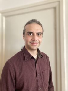 Linus standing in front of a light coloured wall. He wears a maroon button up shirt and his hair is pulled back.