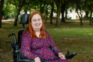 Jess who has long red hair is sitting in her wheelchair in a park of leafy green trees. She's smiling broadly at the camera and is wearing a bright pink and purple leopard print top.
