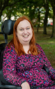 Jess who has long red hair is sitting in her wheelchair in a park of leafy green trees. She's smiling broadly at the camera and is wearing a bright pink and purple leopard print top.