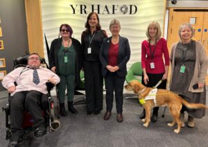 A group photo at the launch of the Disabled People’s Rights Plan. Present are Cabinet Secretary for Social Justice Jane Hutt MS, Disability Rights Taskforce Chair Prof Debbie Foster and Working Group Chairs Damian Bridgeman, Willow Holloway, Andrea Gordon, her guide dog Mae, and Rhian Davies. They're all smiling at the camera.