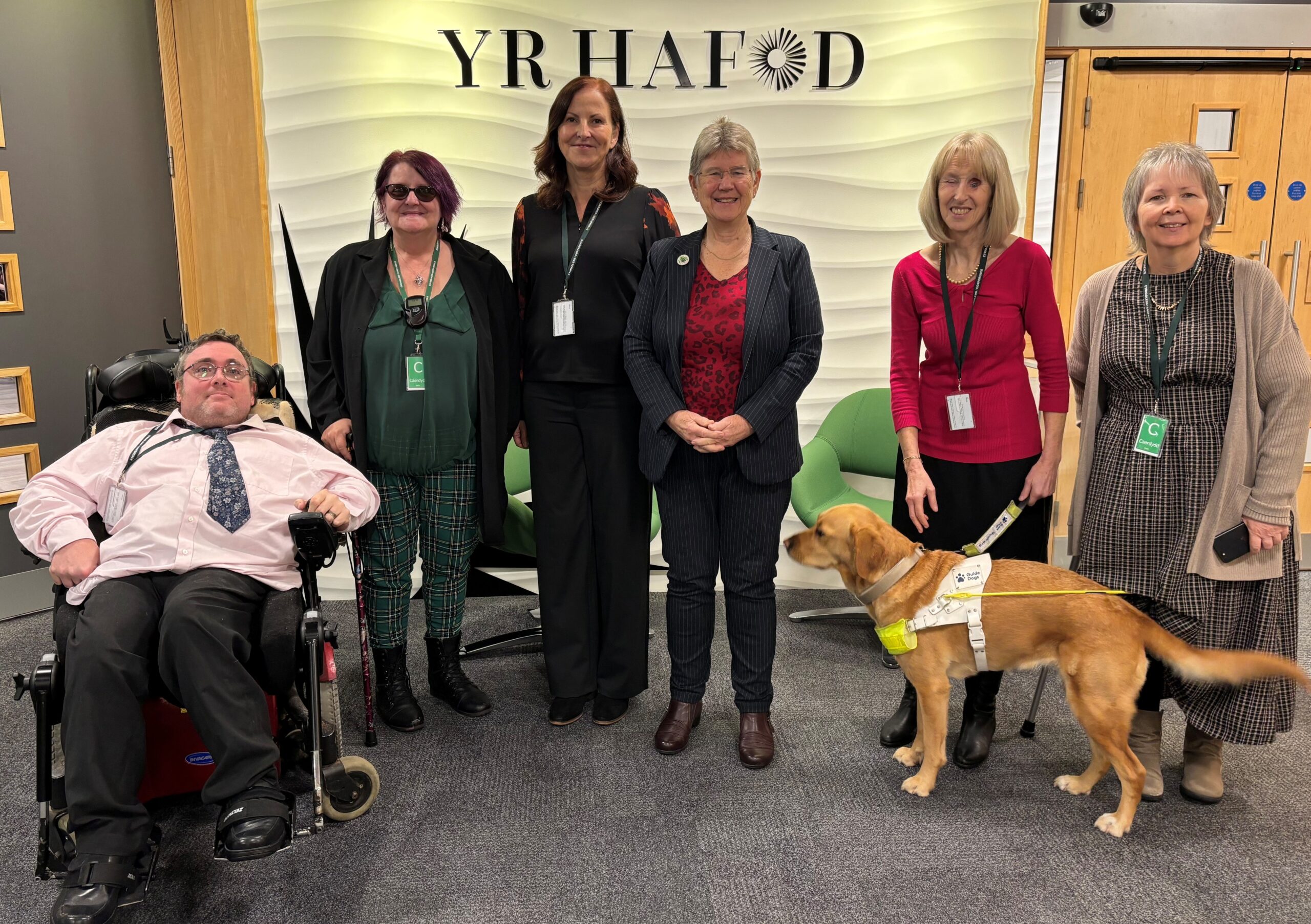 A group photo at the launch of the Disabled People’s Rights Plan. Present are Cabinet Secretary for Social Justice Jane Hutt MS, Disability Rights Taskforce Chair Prof Debbie Foster and Working Group Chairs Damian Bridgeman, Willow Holloway, Andrea Gordon, her guide dog Mae, and Rhian Davies. They're all smiling at the camera.