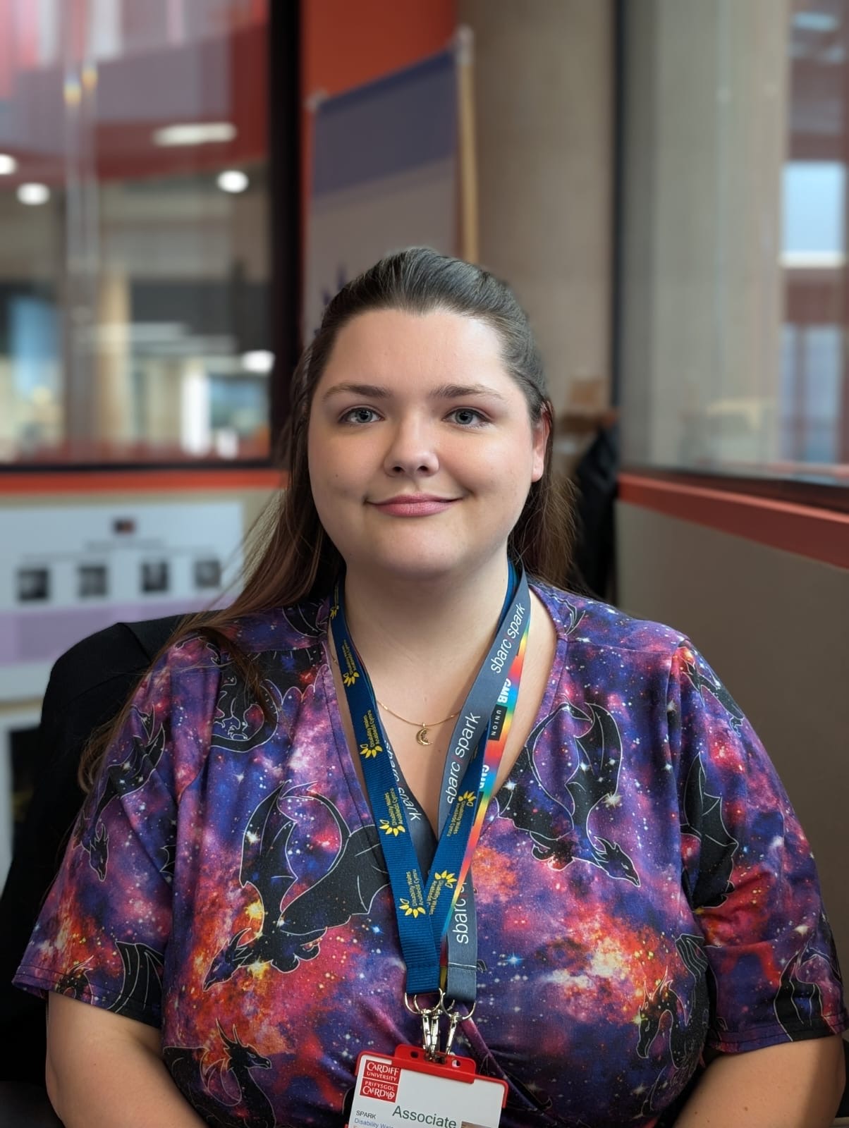 Leandra, who has long brown hair pulled back facing the camera, smiling with long brown hair pulled back. They are wearing a short‑sleeved, multicolour galaxy‑patterned top and lanyards around her neck holding an ID badge.